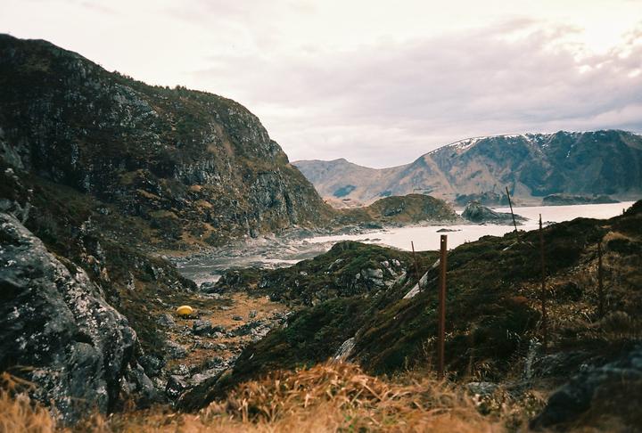 A yellow tent is pitched in a rocky valley leading to a body of water surrounded by mountains.