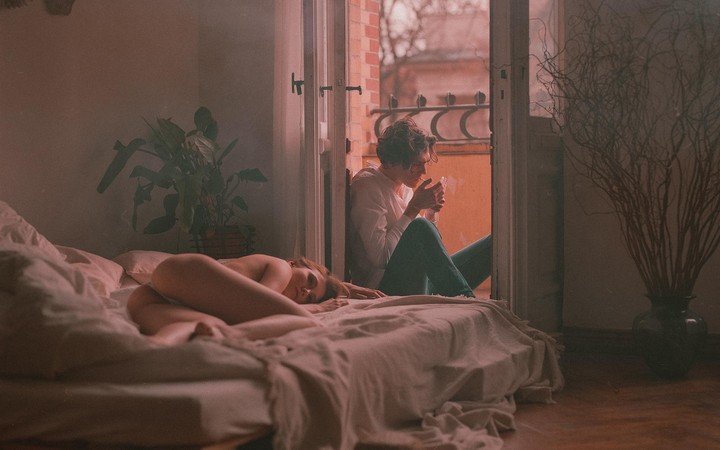 A woman lies in bed while a man sits in a doorway lighting a cigarette.