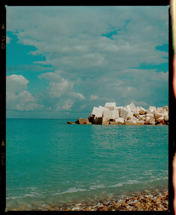 A rocky beach with turquoise water and a breakwater made of concrete blocks under a cloudy sky.