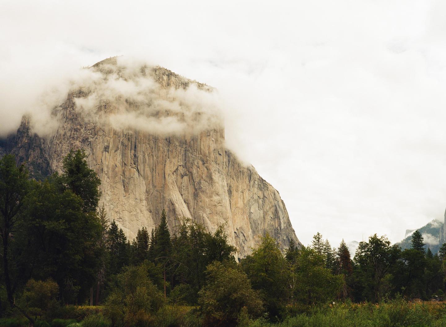 A large rock formation with clouds around its peak rises above a forest.