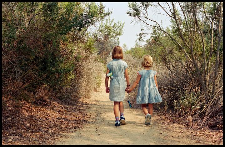 Two girls walk hand in hand down a dirt path surrounded by bushes and trees.