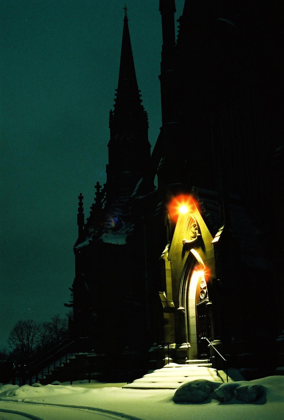 A gothic church with a tall spire is illuminated by a bright light over its entrance, surrounded by snow at night.