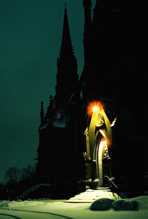 A gothic church with a tall spire is illuminated by a bright light over its entrance, surrounded by snow at night.