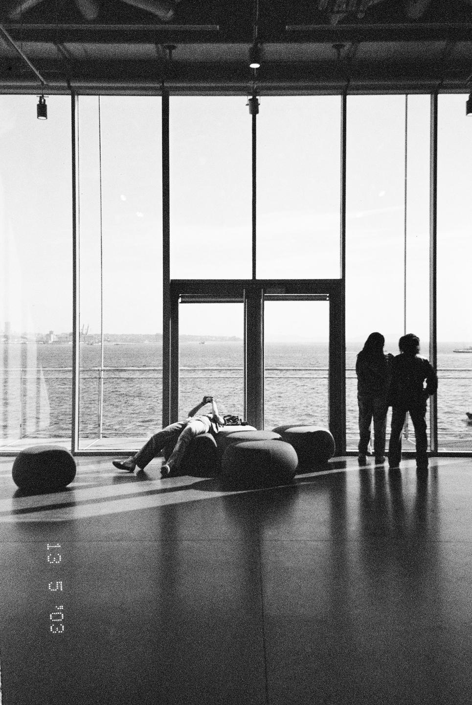 A person reclines on a beanbag while two people stand by a large window overlooking the water.
