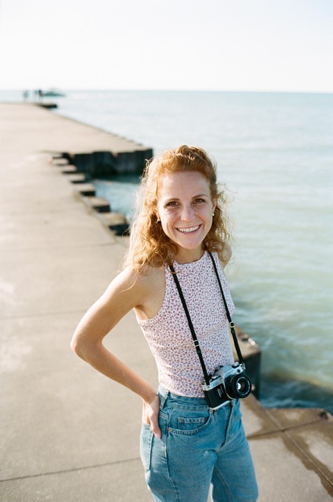 A smiling woman with curly red hair stands on a concrete pier next to a body of water, wearing a floral top, jeans, and a camera around her neck.
