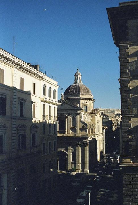A domed church and other buildings line a street with parked cars under a clear sky.