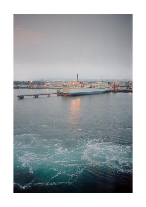 A large ferry is docked at a pier with a town in the background under a cloudy sky, and foamy water is visible in the foreground.