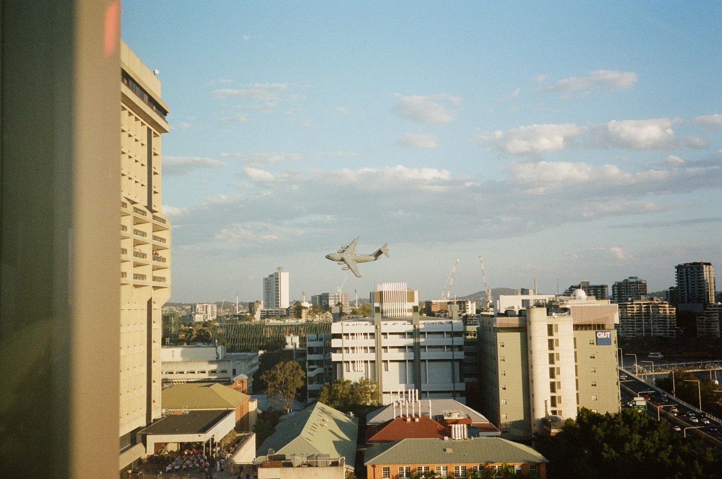 A large airplane flies low over a city with many buildings and a bridge.