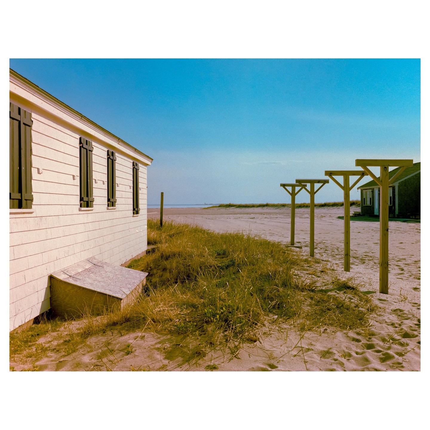 A white house with dark shutters stands on a sandy beach next to tall grass and wooden posts.