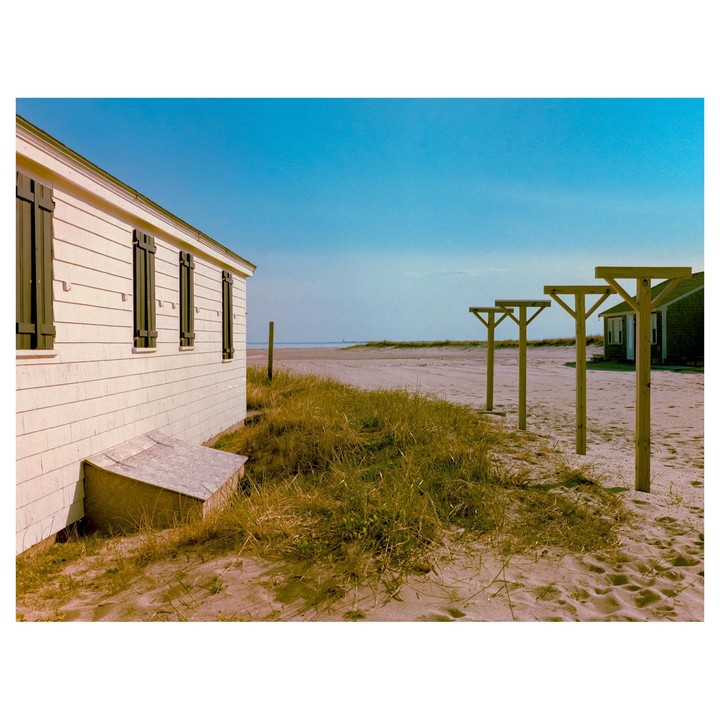 A white house with dark shutters stands on a sandy beach next to tall grass and wooden posts.