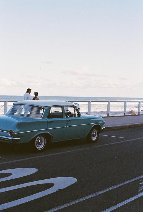 A teal vintage car is parked on a street next to a couple looking out at the ocean.