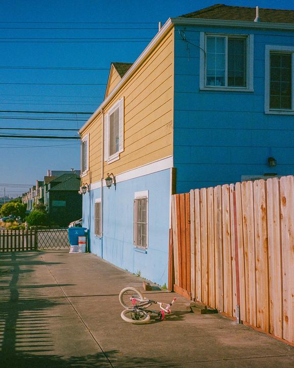 A child's bicycle lies on a concrete driveway next to a blue and yellow building with a wooden fence.