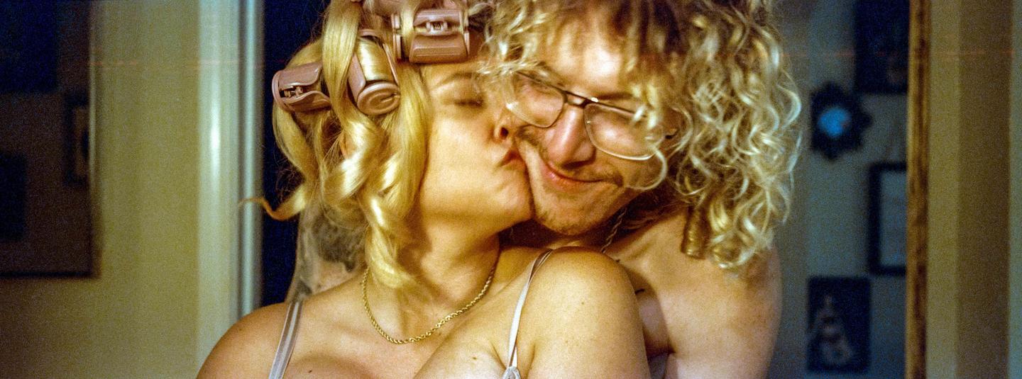 A woman with curlers in her hair kisses a man with glasses and curly hair.