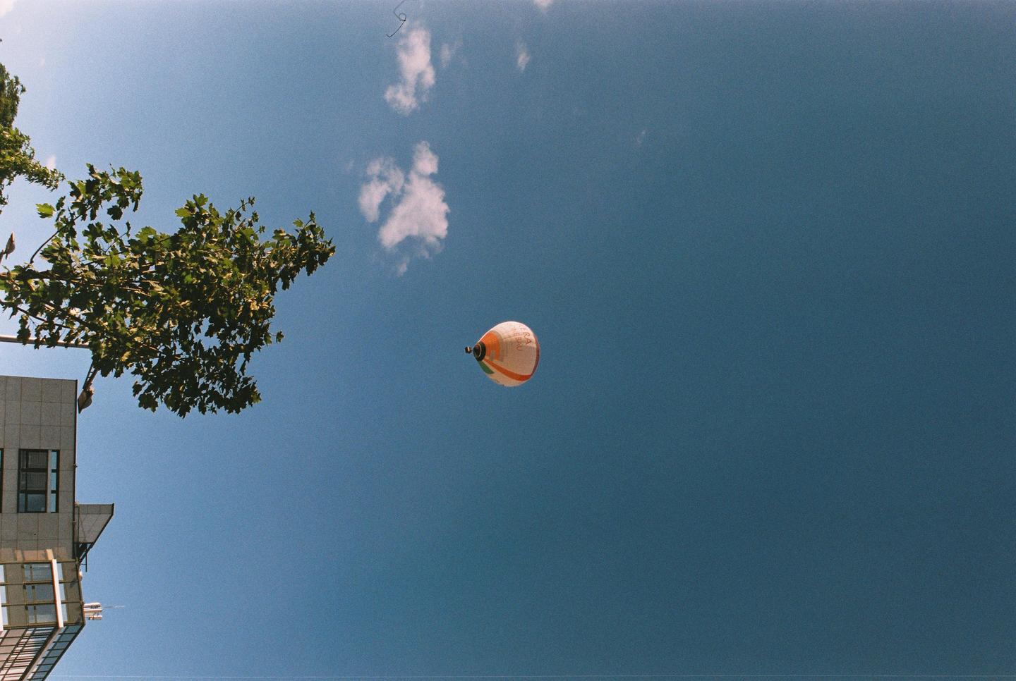 A hot air balloon floats in a clear blue sky above a building and tree.
