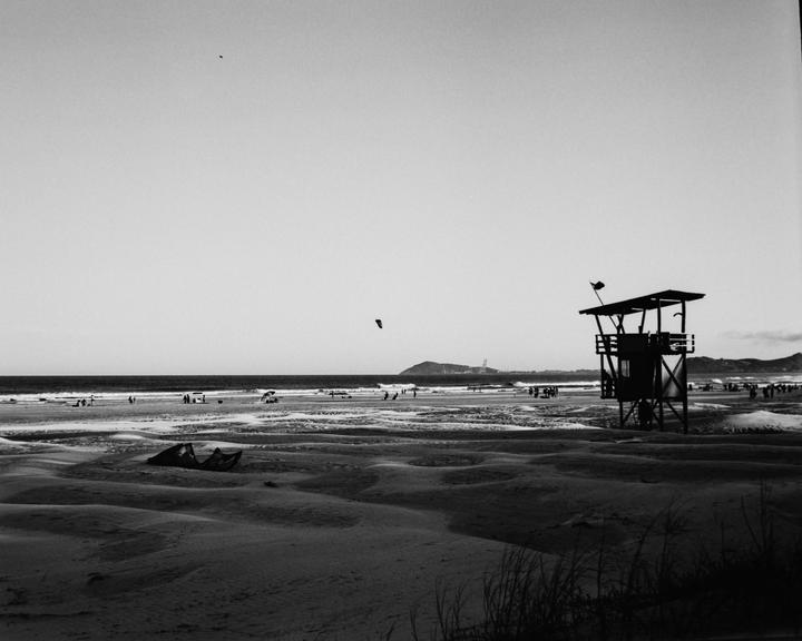 A black and white image shows a beach with a lifeguard tower on the right, people in the distance, and a kite in the sky.