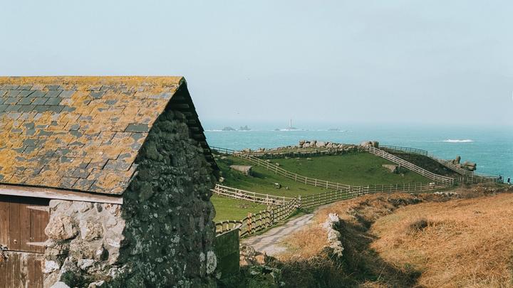 A stone building with a mossy roof stands in the foreground, overlooking a green hillside with fences leading to the ocean and a distant lighthouse.