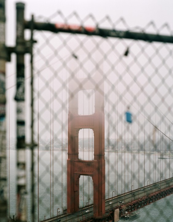 The Golden Gate Bridge is visible through a chain link fence on a foggy morning.