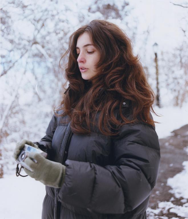 A woman in a black puffer jacket and gloves holds a small camera in a snowy landscape.