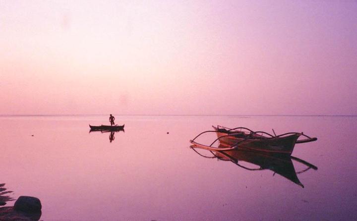 A man stands in a small boat on calm water with two other boats floating nearby at dusk.