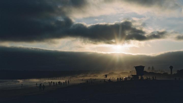The sun shines through clouds over a beach with many people and a lifeguard stand.