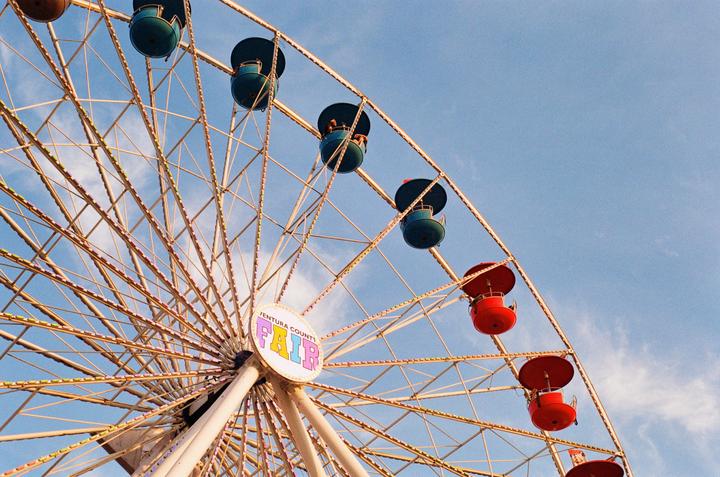 A low-angle view shows a ferris wheel with red and teal gondolas against a blue sky with some clouds.