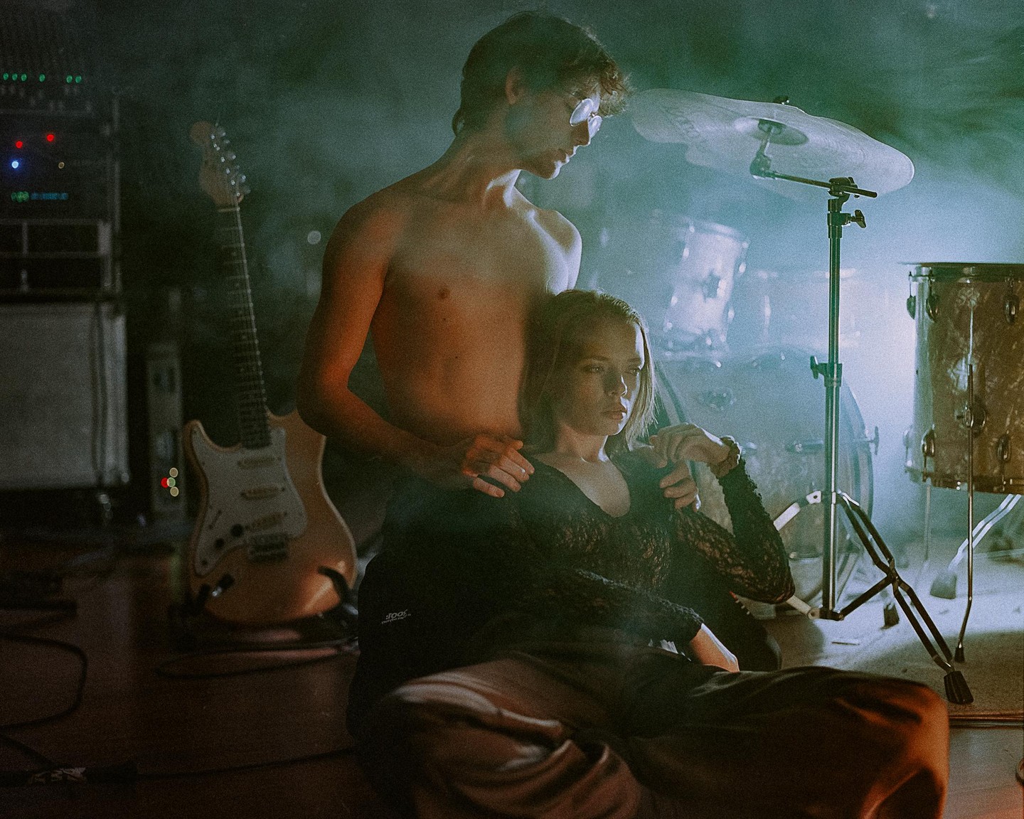 A shirtless man stands behind a woman sitting on the floor, with a guitar and drum set in the background.
