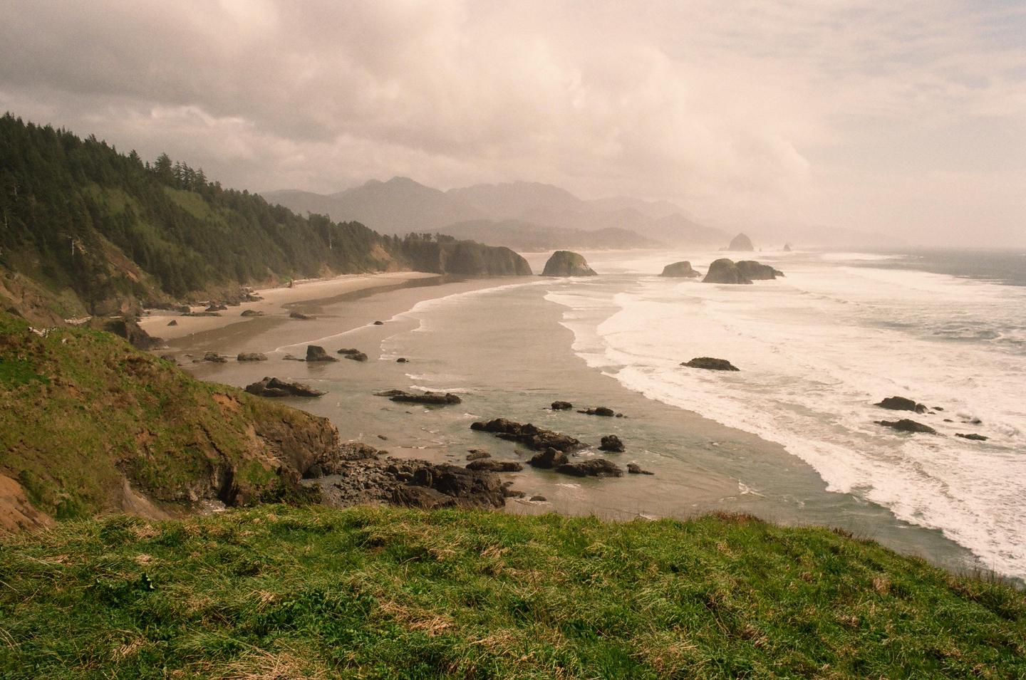 A wide view of the Oregon coast shows a sandy beach, forested hills, and rock formations in the ocean under a cloudy sky.