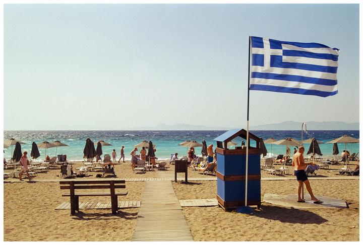 A Greek flag flies over a sandy beach with people, umbrellas, and the sea in the background.