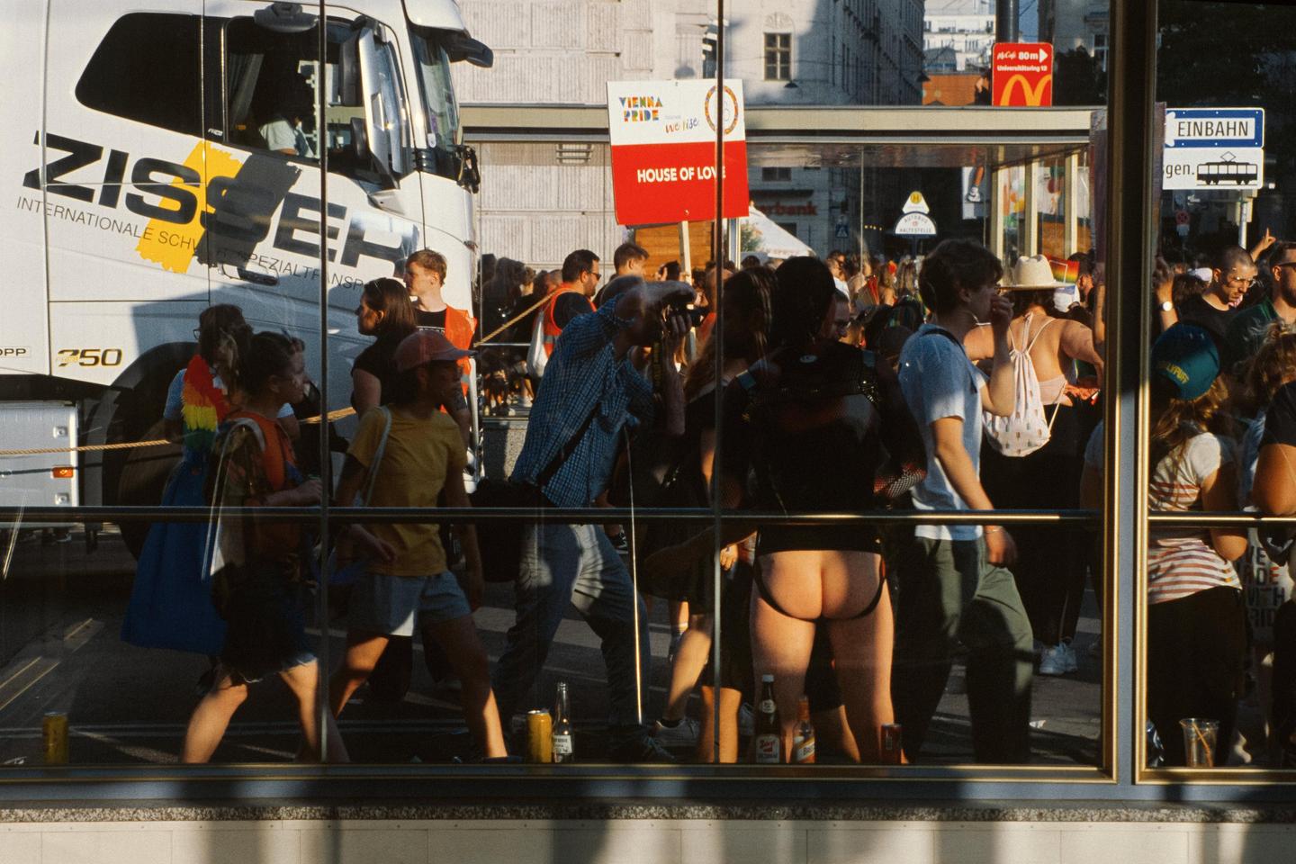 A crowd of people are gathered on a city street, with a large truck and a sign for "Vienna Pride" visible in the background.