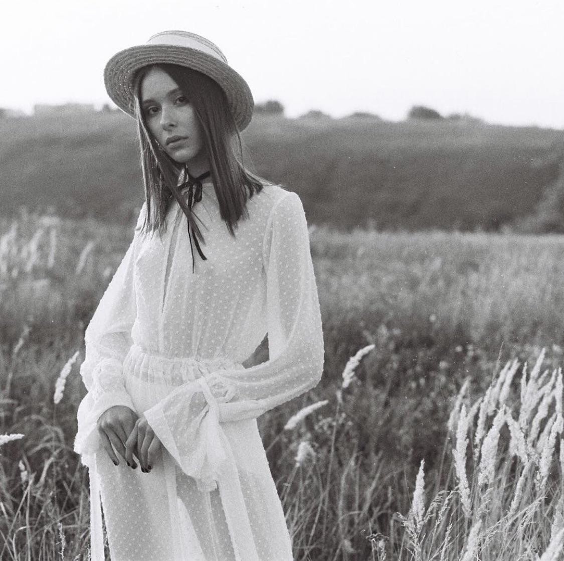 A woman in a white dress and straw hat stands in a field of tall grass.