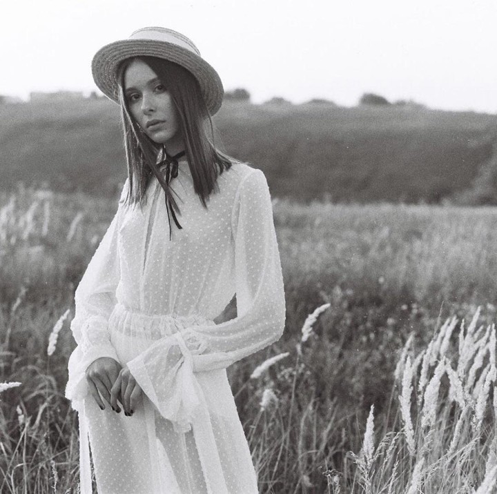 A woman in a white dress and straw hat stands in a field of tall grass.