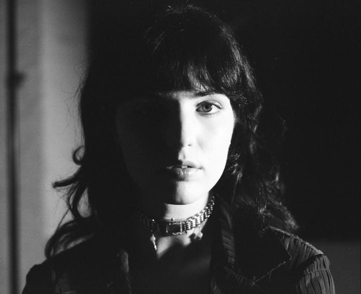 A woman with dark hair and a choker necklace looks directly at the viewer, with half of her face in shadow.