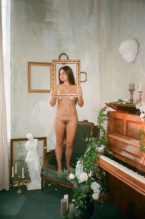 A nude woman stands on a chair holding a picture frame in front of her chest, with a piano and a statue in the background.