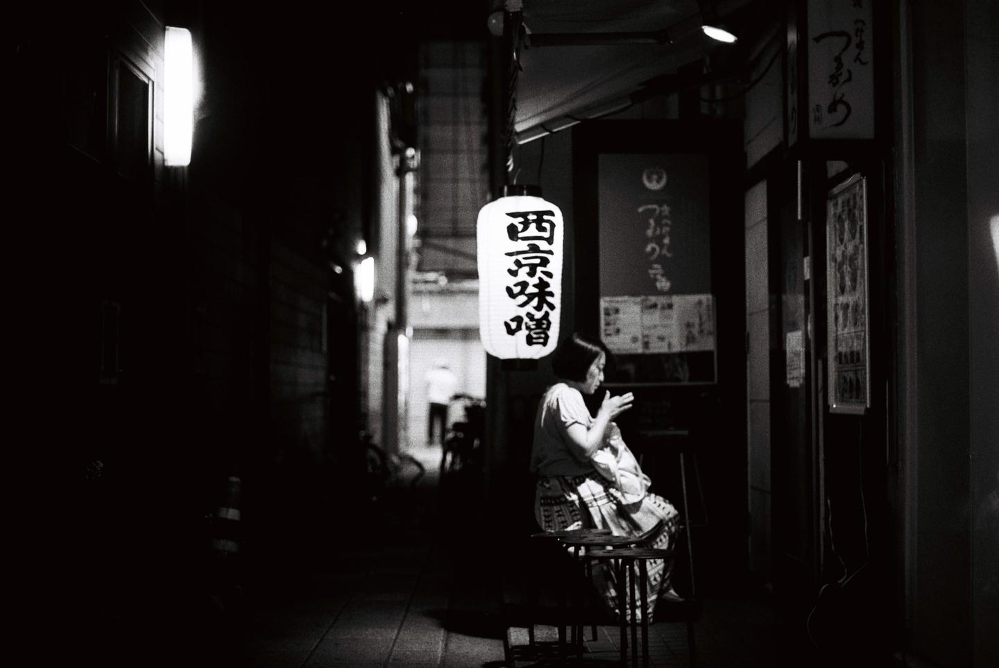 A woman sits on a stool under a hanging lantern on a dark street at night.