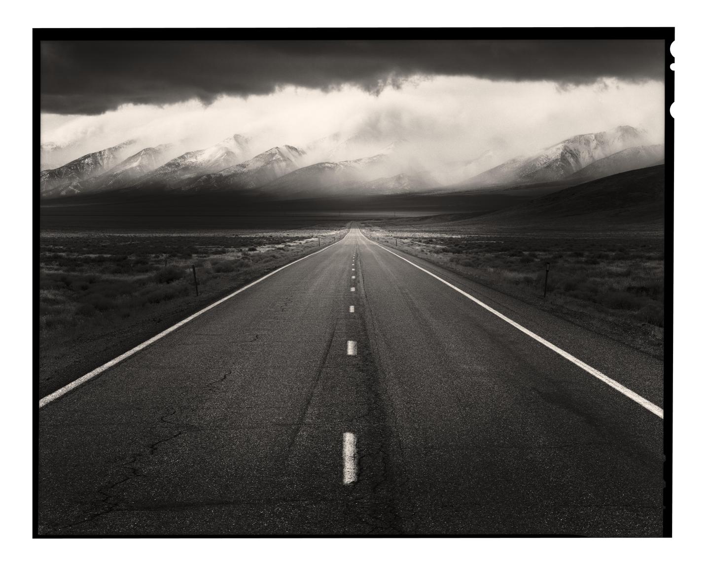 A long, straight road leads towards snow-capped mountains under a dramatic, cloudy sky.