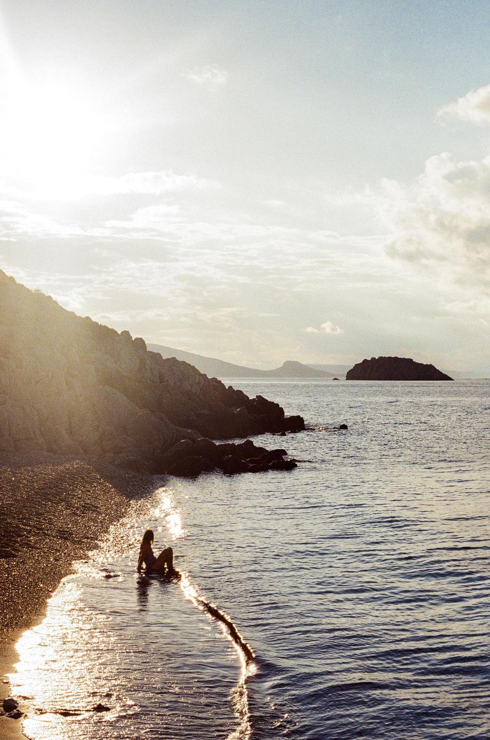 A woman sits on a rocky beach at the water's edge with the sun reflecting on the water.