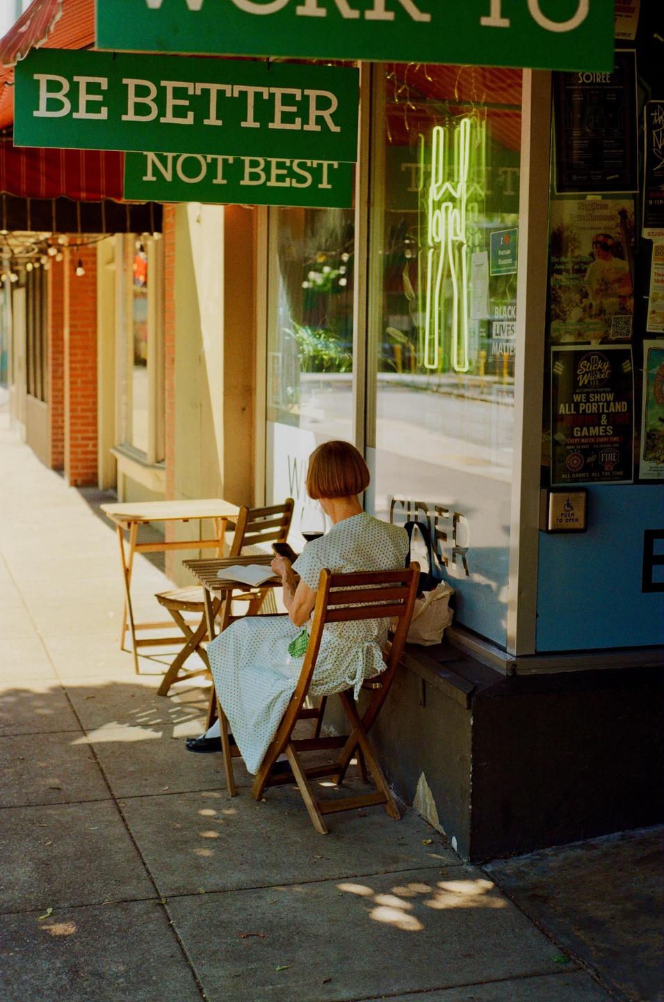 A woman sits at a small table on a sunlit sidewalk next to a storefront with a green awning and neon sign.