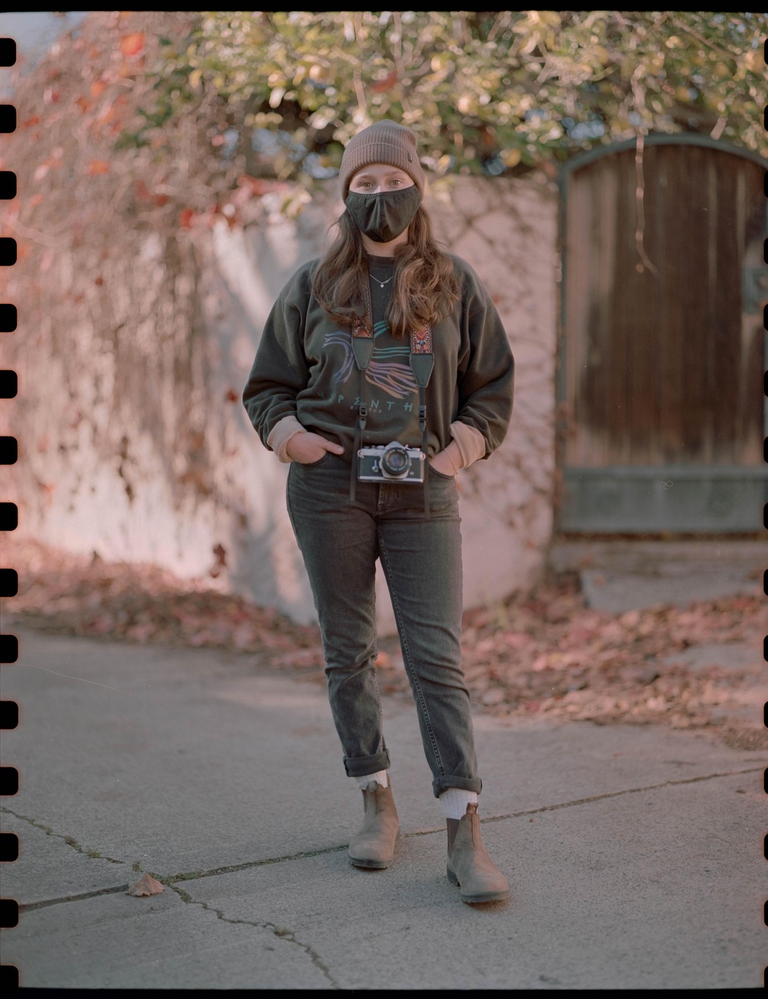 A woman wearing a beanie and a mask stands on a paved surface with a camera around her neck.