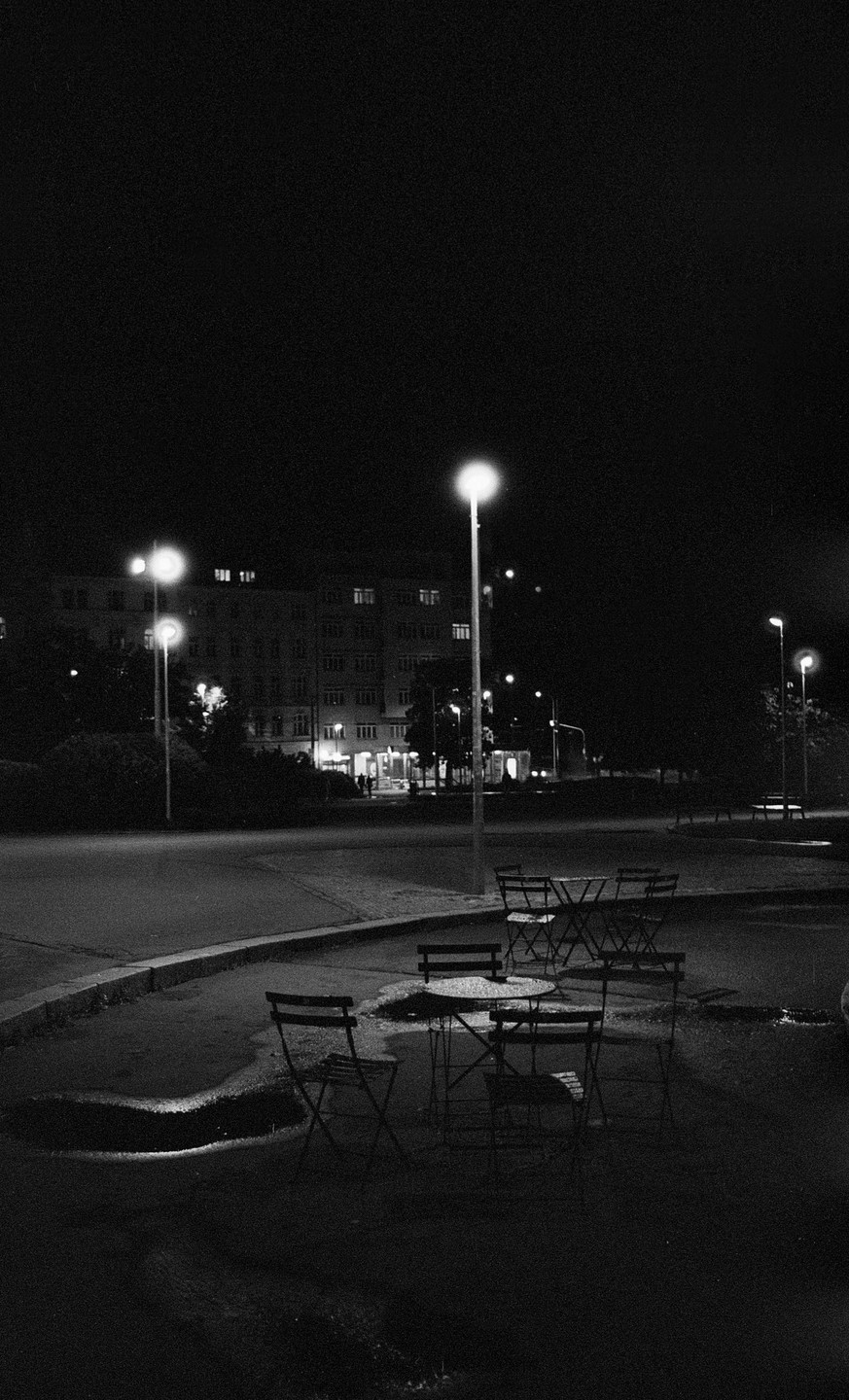 A street at night with tables and chairs in the foreground and a building in the background.
