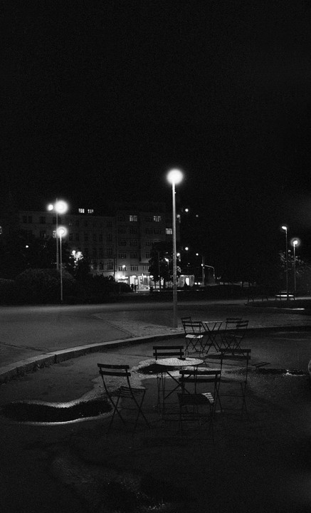 A street at night with tables and chairs in the foreground and a building in the background.