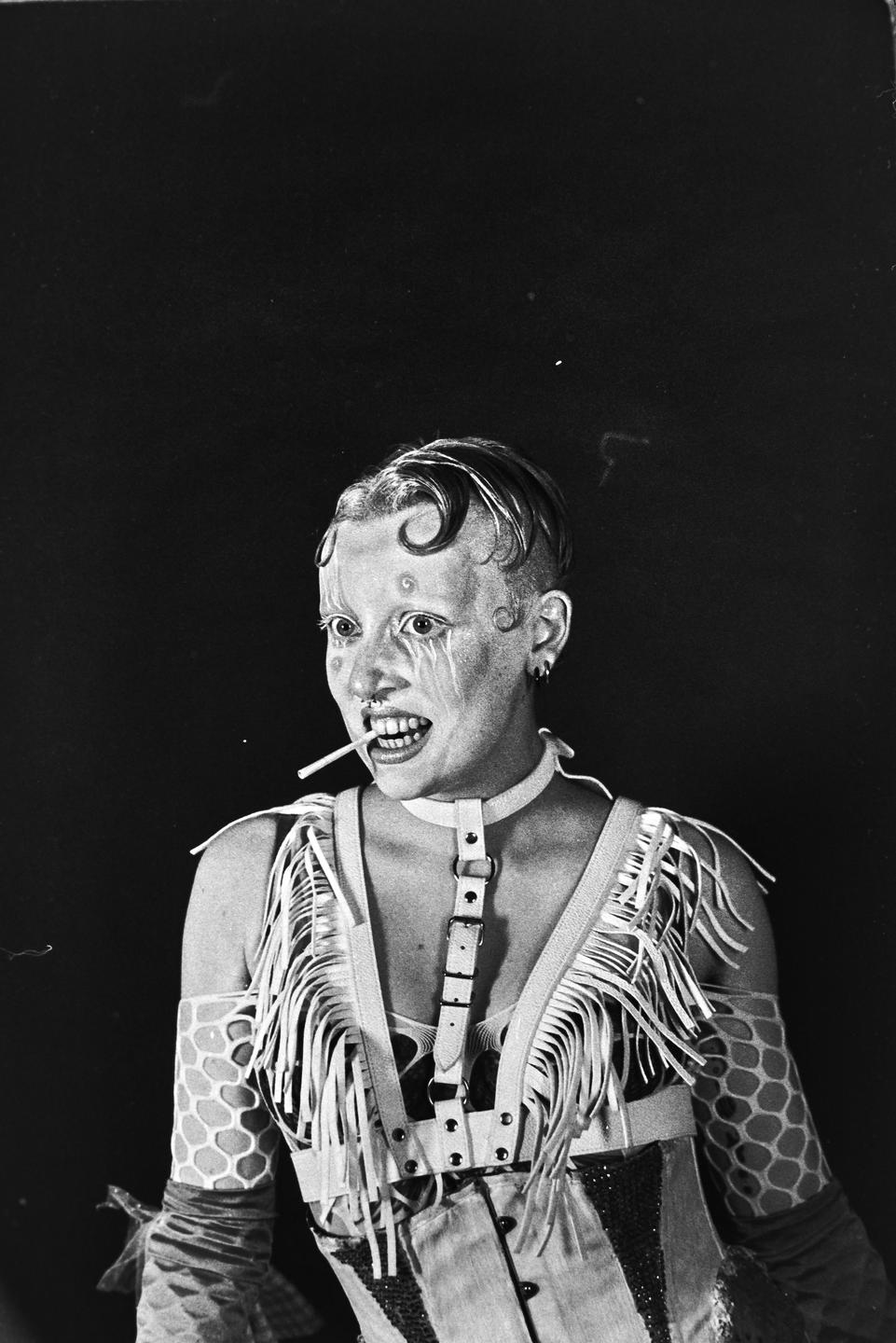 A person with dramatic makeup and an elaborate outfit holds a lollipop in their mouth.