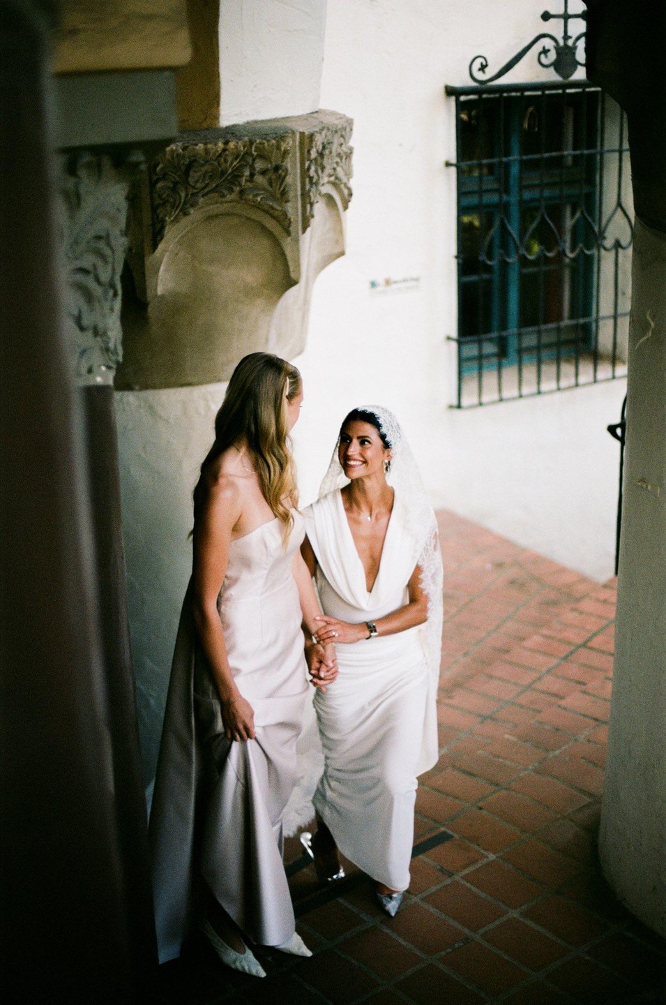 Two brides in wedding dresses hold hands and smile at each other in a hallway with a tiled floor and a window with a wrought iron grate.