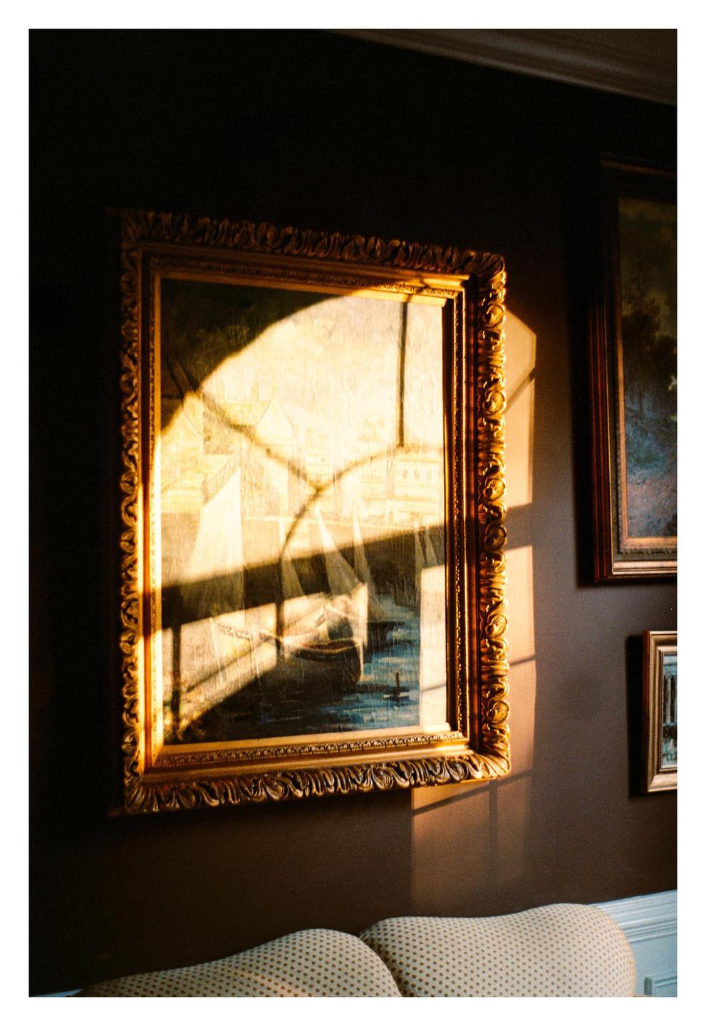 Sunlight streams through a window, casting shadows on a framed painting of boats on the water.