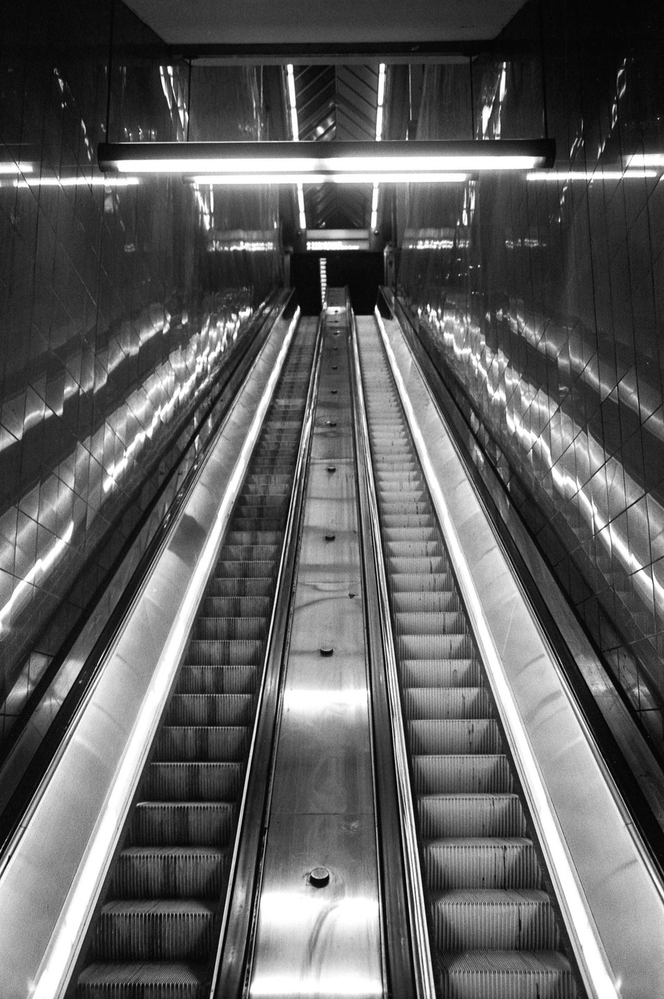 Two long escalators descend into a dark subway station, illuminated by overhead lights and reflections on the tiled walls.