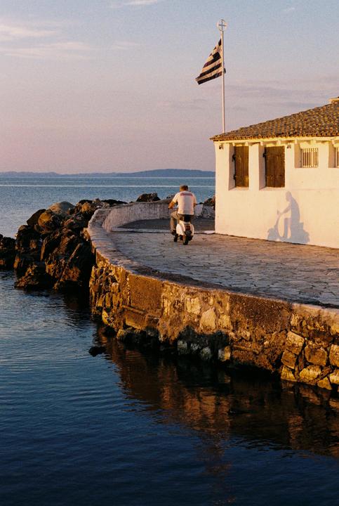 A man rides a scooter along a stone path next to a body of water with a white building and a flag.