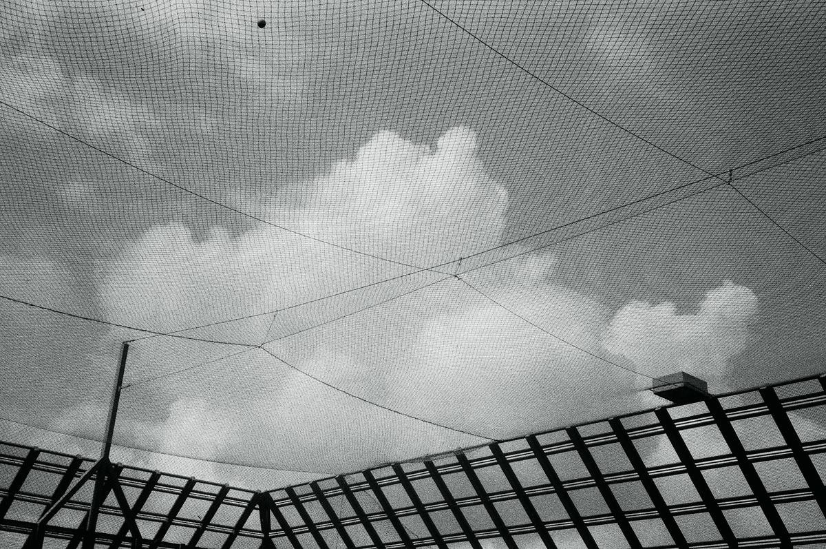 A baseball net covers the sky with clouds and a building visible below.