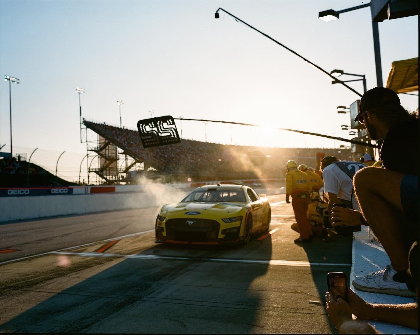 A yellow race car is on a track with people in the pit crew and a man watching from the side.