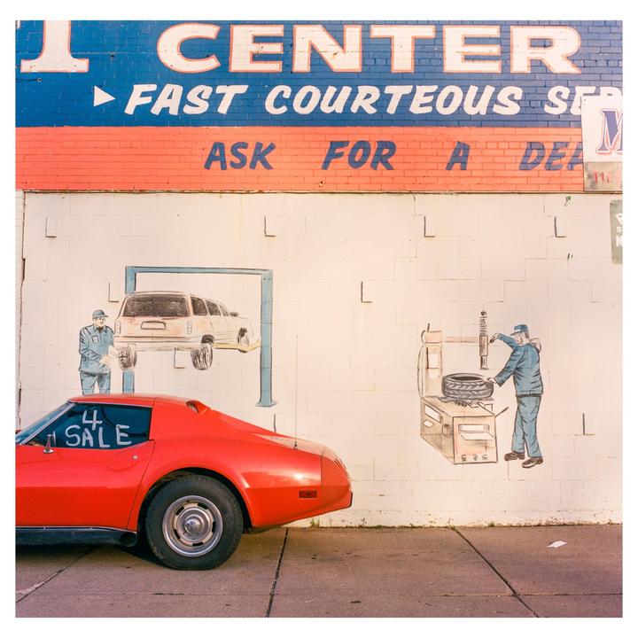 A red car with a "4 Sale" sign in its window is parked in front of a building with a mural of mechanics and cars.