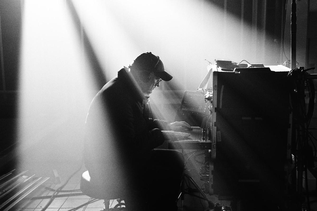 A man wearing a cap and headphones is sitting at a mixing console, illuminated by bright, hazy light.