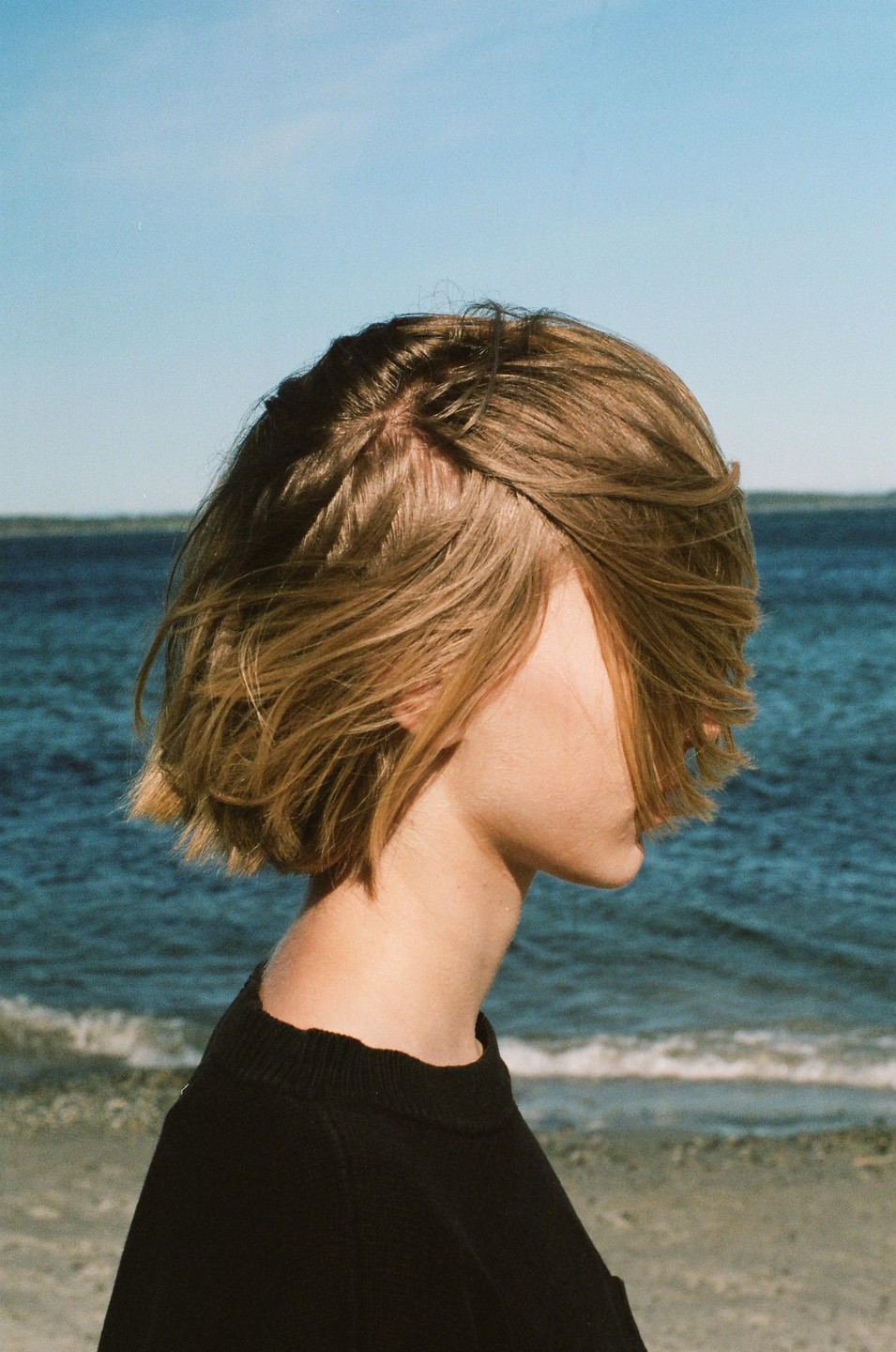 A woman with short hair stands in profile on a beach with the ocean in the background.